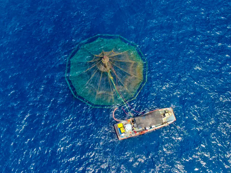 Aerial view of a boat tending an offshore aquaculture net pen in open blue water