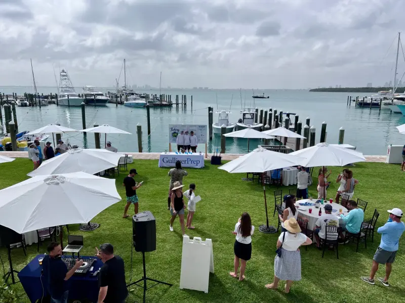 Waterfront lawn at the Big Catch Fishing Tournament in Miami: guests under white umbrellas, a Gulliver Prep table, and boats docked on the bay