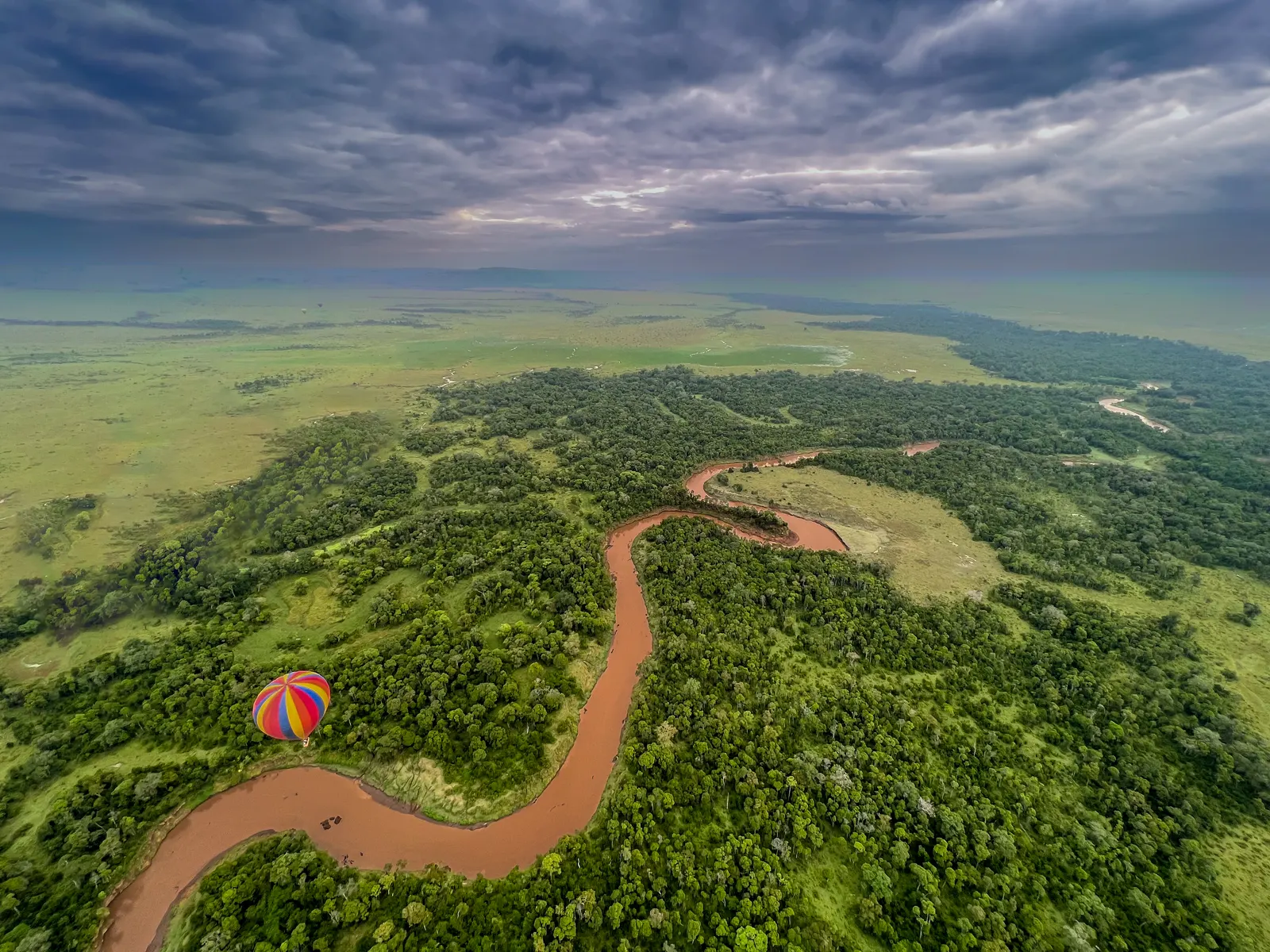 Aerial view of a red river winding through green forest with a hot air balloon below